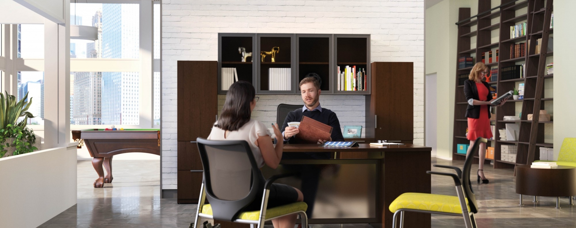 open air office space with a man and woman discussing work at his desk. Yellow stationary high back chairs. Dark wood wall height bookshelves line the walls. 
